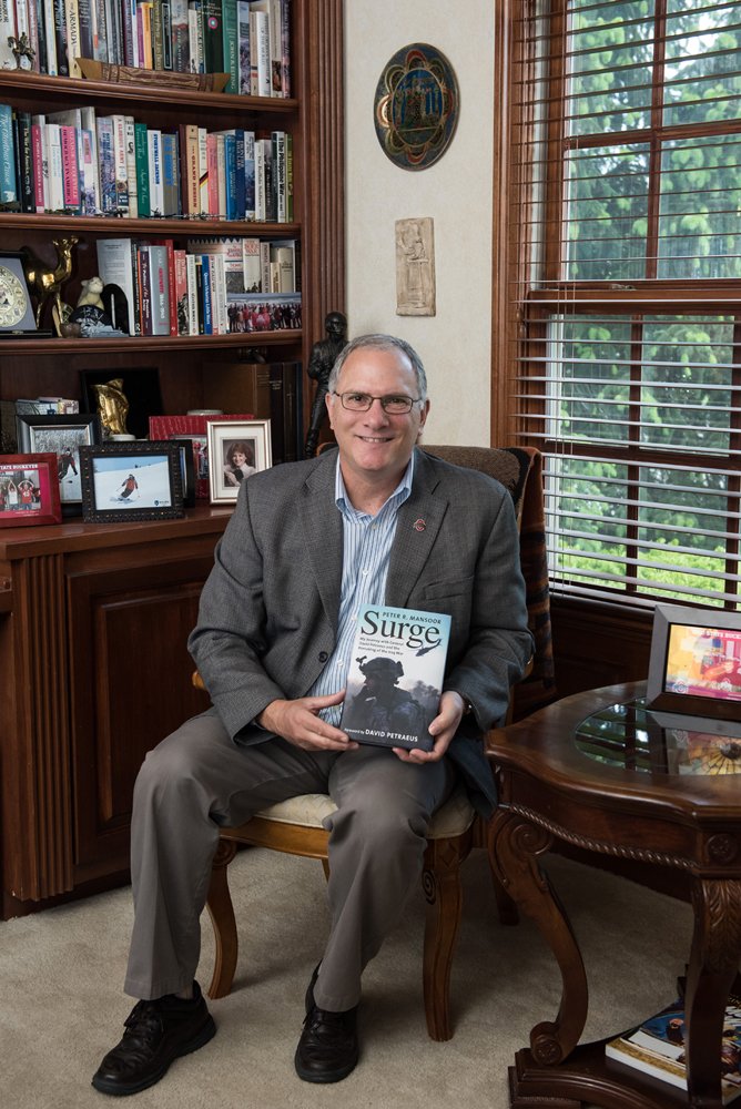 A man in a gray suit sits in a chair in an office holding a book titled "Surge." Photo Credit: Gwendolyn Z. Photography